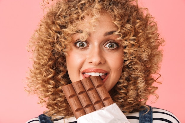 Foto vrolijke gekrulde vrouw 20s die chocoladereep eet terwijl hij staat