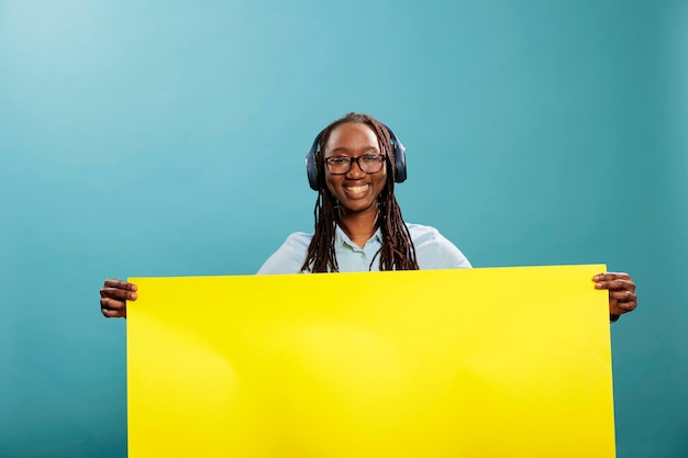 Vrolijke gelukkig lachende zelfverzekerde vrouw met gele blanco papieren bannerbord voor advertentieplaatsing. Vrolijke jonge dame met een leeg reclamebord en draadloze koptelefoon die naar de camera glimlacht