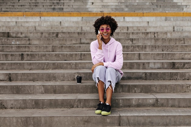 Vrolijke gelukkige krullende vrouw in stijlvolle jeans, oversized hoodie en roze zonnebril glimlacht, praat aan de telefoon en zit buiten op trappen