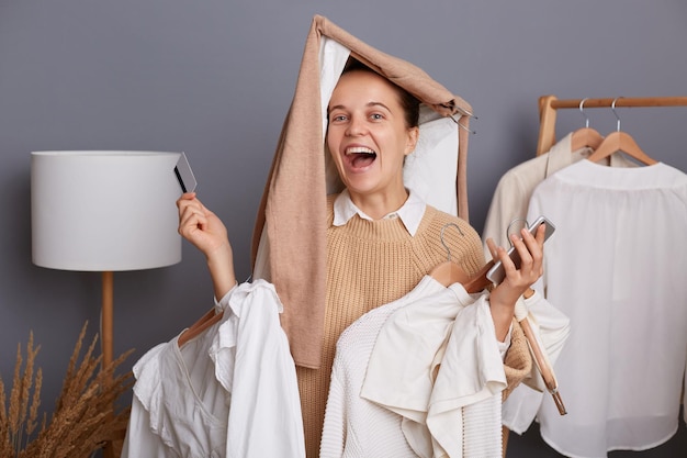 Vrolijke, gelukkige lachende vrouw in een beige trui poserend met telefoon en creditcard die zich verheugt over haar nieuwe aankopen die tegen de grijze muur staan met kleren hangen in kledingkast op rek