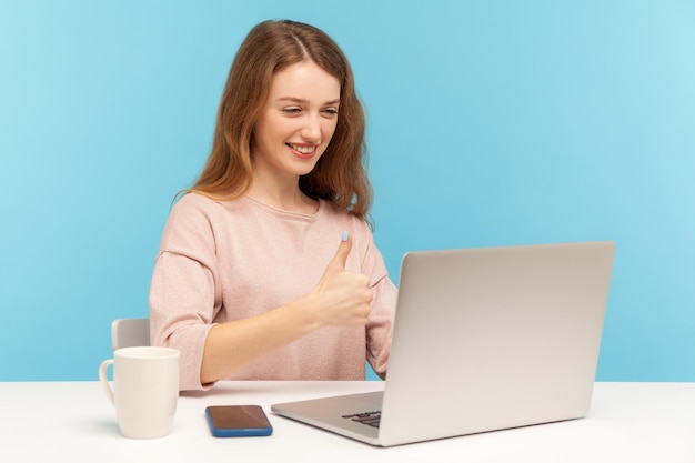 Vrolijke, gelukkige zakenvrouw in vrijetijdskleding die naar het scherm van de laptop kijkt en duimen laat zien, pratend op videogesprek, online conferentie thuiskantoor. indoor studio-opname geïsoleerd op blauwe achtergrond