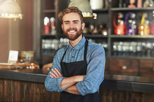 Foto vrolijke glimlach en portret van de eigenaar van de coffeeshop die bij de toonbank staat in zijn startup-cafetaria gelukssucces en mannelijke barista met klein bedrijf met gekruiste armen voor vertrouwen in restaurant