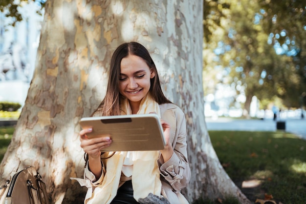 Vrolijke glimlach op het gezicht van een student die onder een boom zit en naar een tablet-pc kijkt