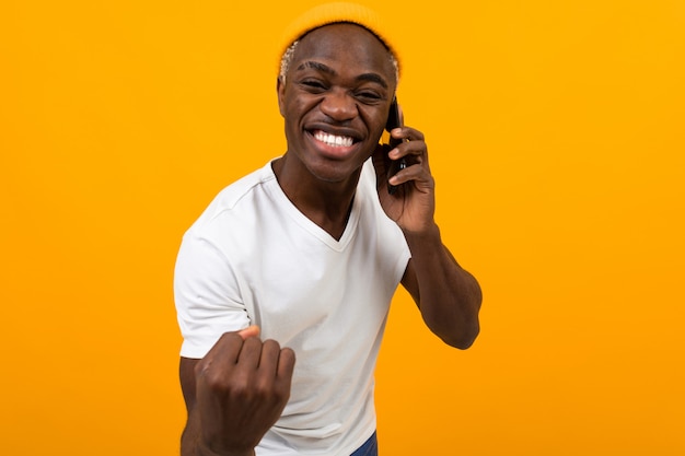 Vrolijke glimlachende Amerikaanse mens in witte t-shirt die op de telefoon op schone geel spreken