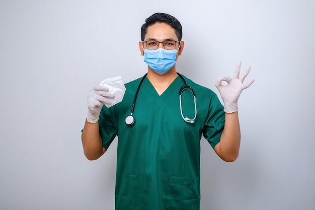 Vrolijke glimlachende aziatische mannelijke arts in scrubs met een medisch masker suggereert een gasmasker terwijl hij een goed teken doet met de vinger