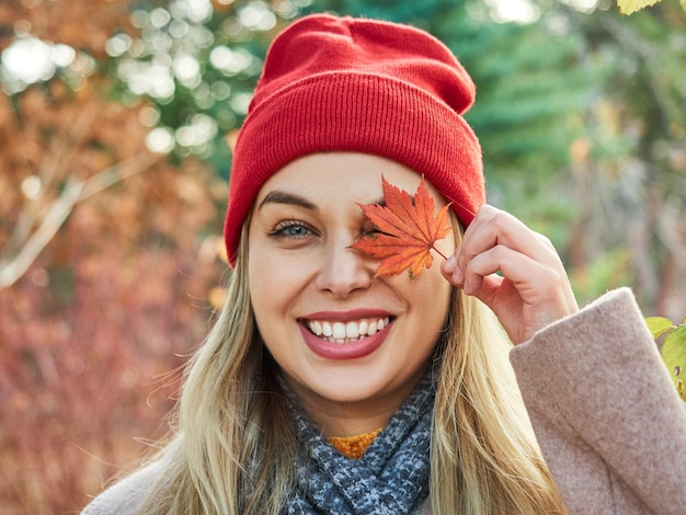 Vrolijke glimlachende blonde vrouw met een rode hoed sluit haar oog met een rood herfstblad