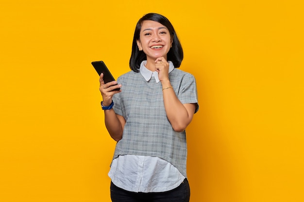 Vrolijke glimlachende jonge Aziatische vrouw met vinger op kin en mobiele telefoon vasthoudend op gele achtergrond