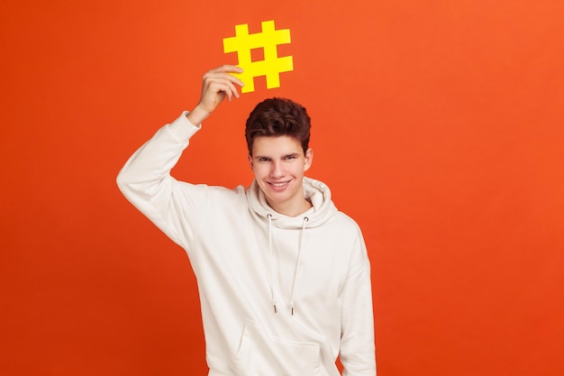 Vrolijke glimlachende jongeman in een casual witte hoodie met een hashtag-teken boven het hoofd, een populaire blogger die internettrends aanbeveelt. Indoor studio-opname geïsoleerd op oranje achtergrond