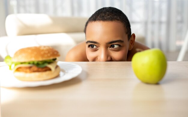 Foto vrolijke grappige jonge afro-amerikaanse dame kijkt naar hamburger aan tafel met groene appel