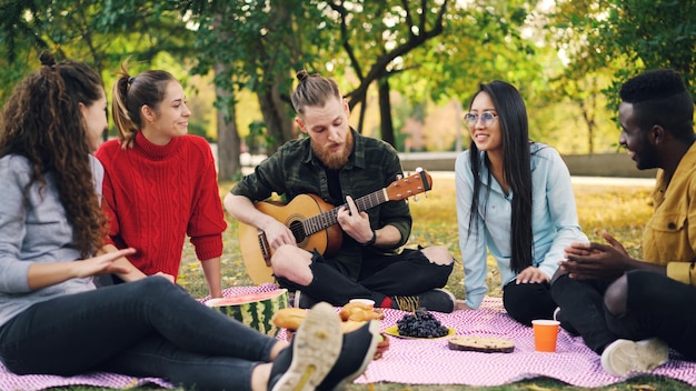 Vrolijke hipster zingt en speelt gitaar, zittend op een deken in het park met vrienden en plezier mensen klappen in de handen Voedselmuziek en leuk concept