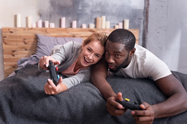 Vrolijke internationale jonge paar spelen van videogames terwijl ze in bed liggen en plezier hebben.