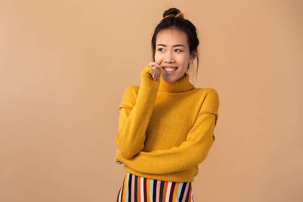 vrolijke japanse vrouw die een trui draagt die lacht en opzij kijkt, geïsoleerd over een beige muur in de studio