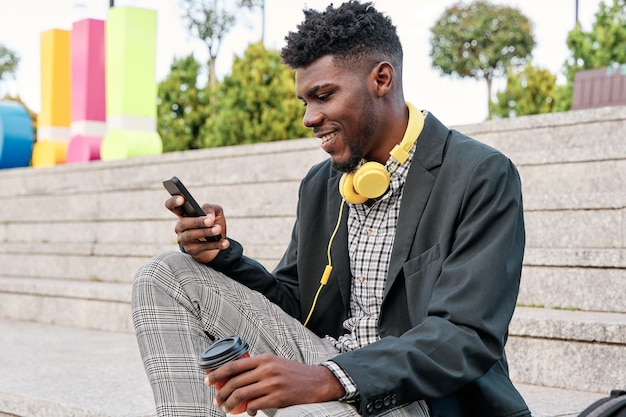 Vrolijke jonge afrikaanse man met blazer buitenshuis met afhaalkoffiekopje met mobiele telefoon