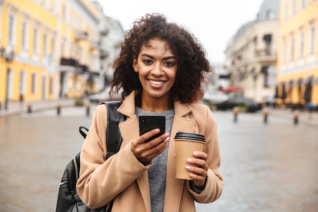 Vrolijke jonge afrikaanse vrouw die jas draagt die buiten loopt, afhaalmaaltijdenkoffie houdt, die mobiele telefoon gebruikt