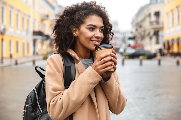 Vrolijke jonge Afrikaanse vrouw die jas draagt die buiten loopt, afhaalmaaltijdenkoffie houdt