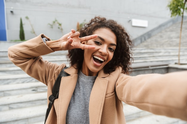 Vrolijke jonge Afrikaanse vrouw die jas draagt die buiten loopt, een selfie neemt