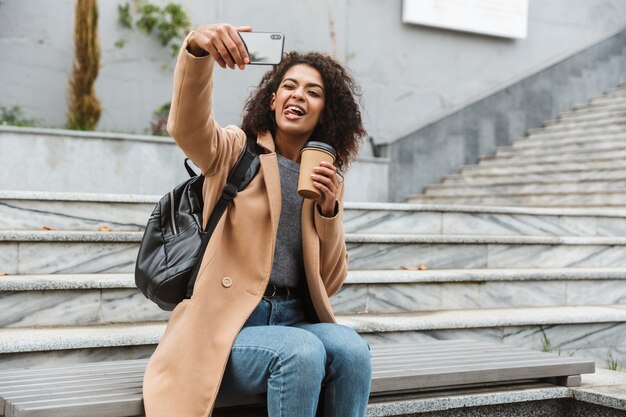 Vrolijke jonge Afrikaanse vrouw die jas draagt die buiten zit, afhaalmaaltijdenkoffie houdt, die een selfie neemt