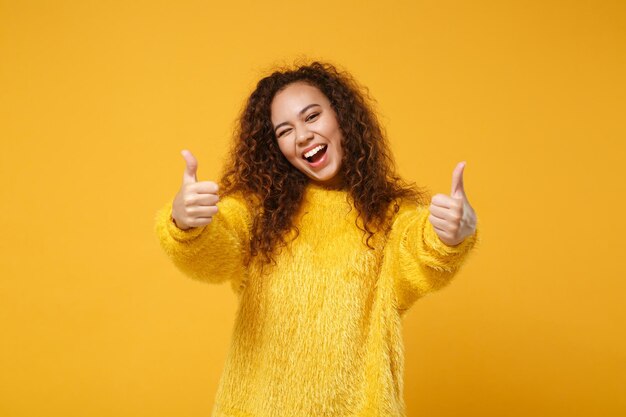 Vrolijke jonge Afro-Amerikaanse meisje in bont trui poseren geïsoleerd op geel oranje achtergrond in studio. Mensen oprechte emoties levensstijl concept. Bespotten kopie ruimte. Met duimen omhoog, knipperend.
