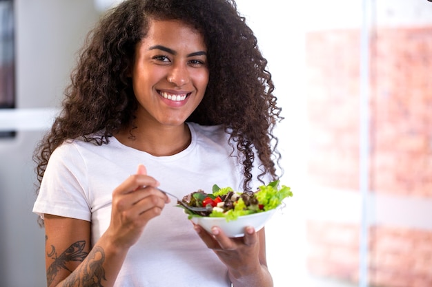 Vrolijke jonge afro-amerikaanse vrouw die groentesalade in huiskeuken eet