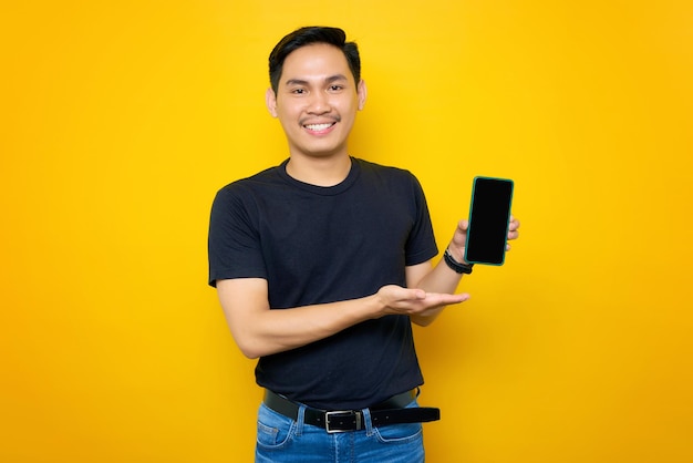 Vrolijke jonge Aziatische man in casual tshirt presenteren mobiele telefoon met leeg scherm geïsoleerd op gele achtergrond Mensen lifestyle concept