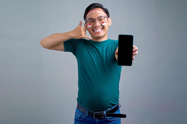 Vrolijke jonge Aziatische man in casual tshirt weergegeven: mobiele telefoon met leeg scherm weergegeven: bel me gebaar geïsoleerd op grijze achtergrond Indonesische onafhankelijkheidsdag viering concept