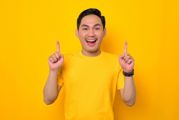 Vrolijke jonge aziatische man in casual tshirt wijzende vinger omhoog met kopie ruimte geïsoleerd op gele achtergrond mensen lifestyle concept