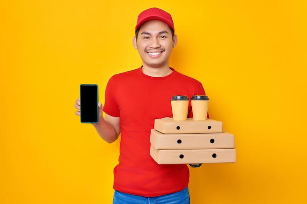 Vrolijke jonge Aziatische man in rode dop tshirt uniforme bezorger werk als dealer koerier houdt pizzadoos en koffie met leeg scherm mobiele telefoon op gele achtergrond Pizza bezorgconcept