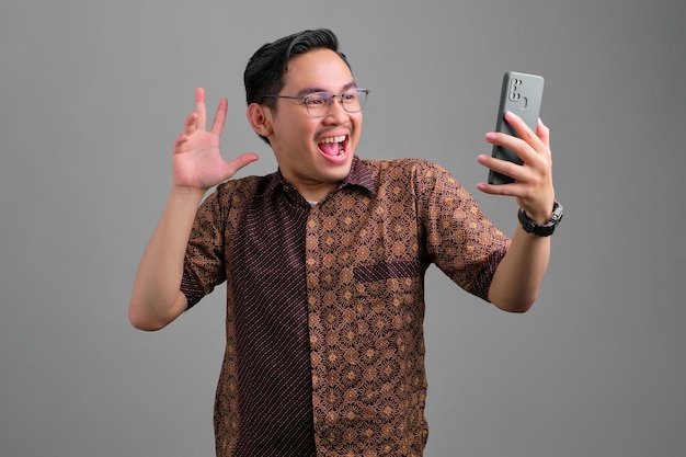 Vrolijke jonge Aziatische man met batik shirt kijken naar smartphone met opwinding en het opsteken van de hand geïsoleerd op een grijze achtergrond