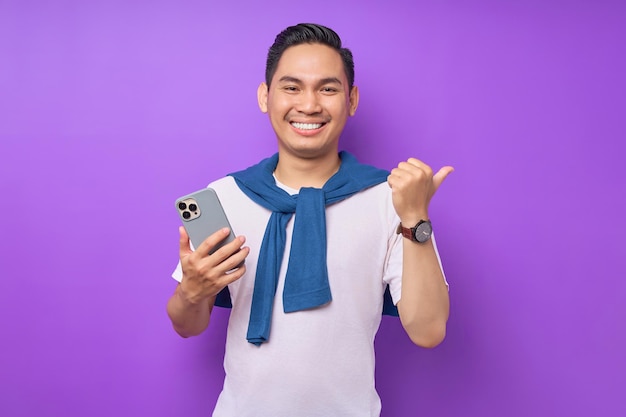 Vrolijke jonge Aziatische man met behulp van mobiele telefoon en duim omhoog gebaar geïsoleerd op paarse achtergrond tonen