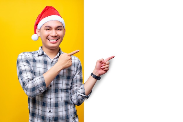 Vrolijke jonge Aziatische man met een kerstmuts staand en wijzend met zijn vinger uit de zijkant van een wit reclamebord geïsoleerd op gele achtergrond Gelukkig Nieuwjaar 2023 vakantieconcept