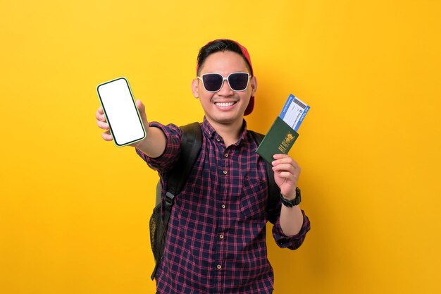 Vrolijke jonge Aziatische man met rugzak met leeg scherm van de mobiele telefoon met paspoort en vliegtickets geïsoleerd op gele achtergrond Toerisme en vakantiereis concept