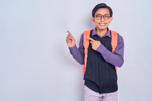 Vrolijke jonge Aziatische mannelijke student van college in casual kleding met rugzak wijzend op iets met vinger geïsoleerd op grijze achtergrond Onderwijs in middelbare school universiteit college concept