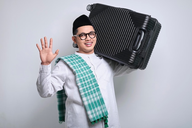 Vrolijke jonge Aziatische moslim man die een koffer op zijn schouder draagt en naar de camera zwaait Ramadan en eid Mubarak concept
