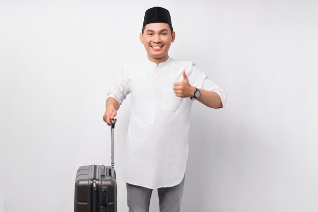 Vrolijke jonge Aziatische moslim man staande terwijl hij koffer in de hand houdt met duim omhoog gebaar klaar voor de vakantie eid Mubarak geïsoleerd op witte achtergrond Ramadan en eid Mubarak concept