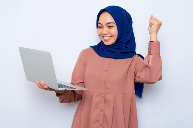 Vrolijke jonge Aziatische moslimvrouw in roze shirt met laptop vieren succes geïsoleerd op witte achtergrond Mensen religieuze levensstijl concept