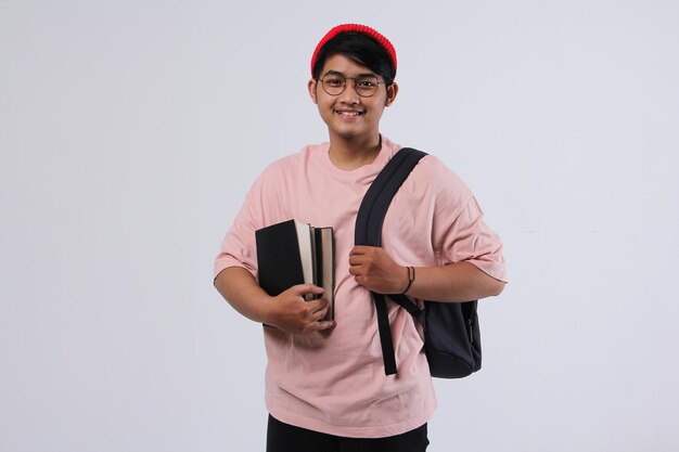 Vrolijke jonge Aziatische universiteitsstudent die staat terwijl hij een boek vasthoudt en een rugzak draagt