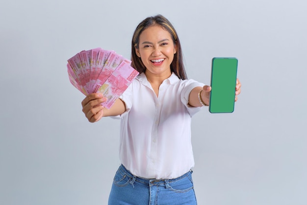 Vrolijke jonge Aziatische vrouw die een lege mobiele telefoon op het scherm toont en geldbankbiljetten vasthoudt die op een witte achtergrond worden geïsoleerd