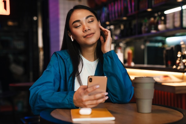 vrolijke jonge aziatische vrouw die mobiele telefoon en draadloze oortelefoons gebruikt terwijl ze in café zit