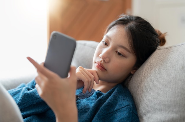 Vrolijke jonge aziatische vrouw die mobiele telefoon gebruikt terwijl ze thuis op een bank zit