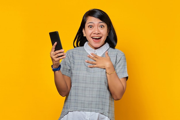 Vrolijke jonge Aziatische vrouw die mobiele telefoon vasthoudt en handpalmen op de borst zet over gele achtergrond