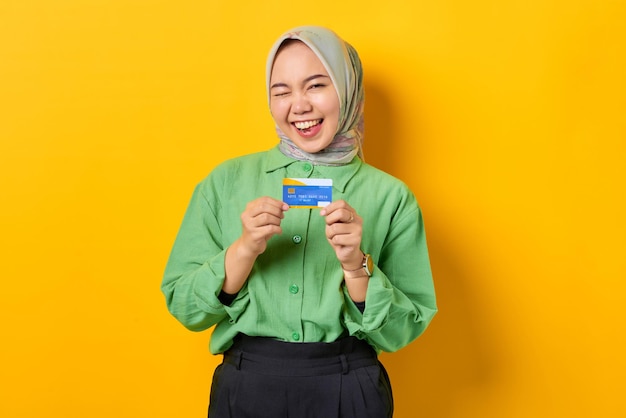 Vrolijke jonge Aziatische vrouw in een groen shirt met creditcard terwijl ze met ogen knipoogt op gele achtergrond