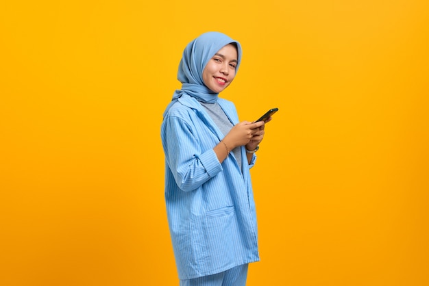 Vrolijke jonge Aziatische vrouw met behulp van mobiele telefoon en kijken naar camera geïsoleerd over gele achtergrond