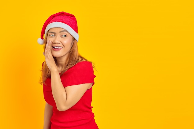 Vrolijke jonge Aziatische vrouw met een kerstmuts die iets fluistert op een gele achtergrond