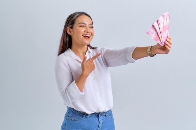 Vrolijke jonge Aziatische vrouw wijzende vinger op geld bankbiljetten geïsoleerd op witte achtergrond