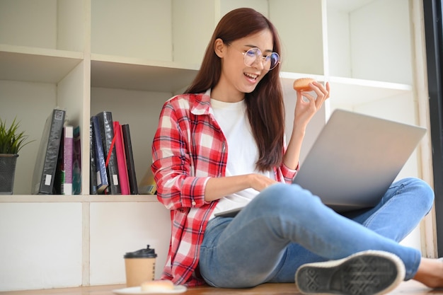 Vrolijke jonge Aziatische vrouwelijke student die draagbare laptop gebruikt die donut eet