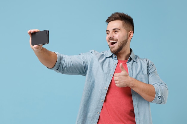 Vrolijke jonge bebaarde man 20s in casual shirt poseren geïsoleerd op pastel blauwe achtergrond in studio. Mensen levensstijl concept. Bespotten kopie ruimte. Selfie geschoten op mobiele telefoon doen, duim opdagen.