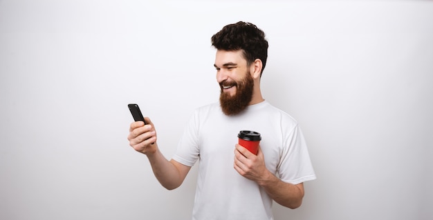 Vrolijke jonge bebaarde man surfen op het internet op zijn telefoon en met een rode papieren beker.