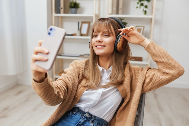 Vrolijke jonge blonde dame in warme trui in koptelefoon luisteren favoriete liedjes doen selfie videogesprek zittend in fauteuil in modern interieur Muziektijd Ontspannen Cool playlist Concept Kopieer ruimte