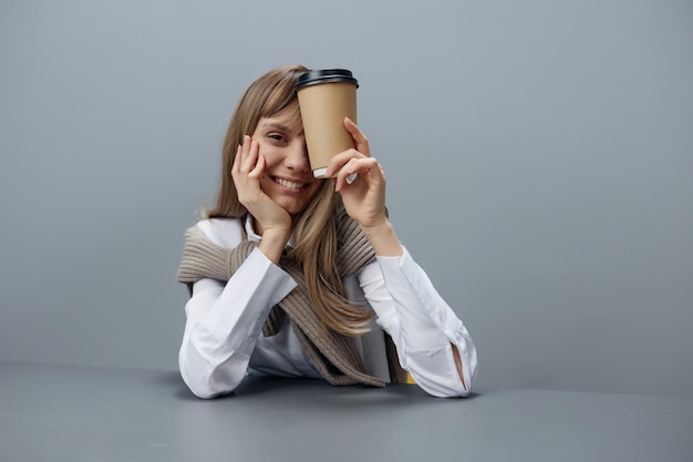 Vrolijke jonge blonde student dame freelancer in warme trui met afhaalkoffie glimlach op camera zittend op de werkplek in grijs modern kantoor aan huis Koffiepauze minnaar Concept kopieer ruimte Cool aanbod