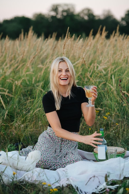 Vrolijke jonge blonde vrouw in een veld die een glas witte wijn optilt